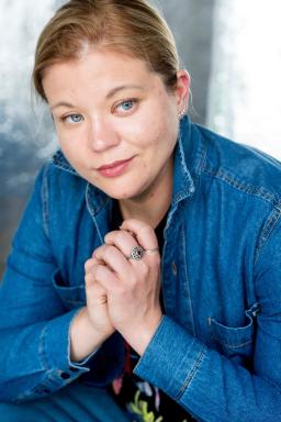 Geneva Halverson, a fair-haired, blue-eyed person, leans toward the camera and is wearing a blue denim shirt.