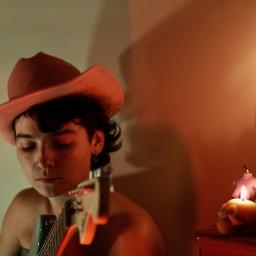 A musician strums a guitar. Her dark hair curls from under a felt brown cowboy hat; her shoulders are bare but her nose and ears are pierced. To the side, a candle shaped like a skull burns beside an unlit pink candle shaped like a woman’s torso.