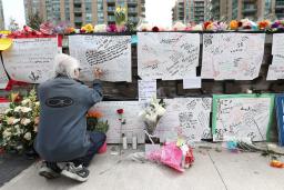 Handmade posters covered with handwritten messages are attached to a low stone wall. A person with white hair and wearing athletic clothes crouches down, writing on a poster.