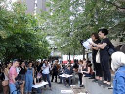 A crowd of a few dozen people gathered in a small urban park listening to two people speaking. The speakers stand on a low wall, holding papers. Some members of the crowd are drawing on protest signs.