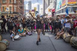 A diverse crowd of people stand in a downtown street, surrounding a Black woman speaking through a megaphone.