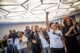 A conference room filled with many Black people of various genders and ages, cheering and smiling enthusiastically, with some raising fists in the air and waving. 