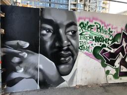 Large graffiti mural consisting of a white, grey and black portrait of Martin Luther King’s face next to stylized green text inside a ragged pink outline, reading “Police Brutality ENDS NOW! RIP Jamal.” 