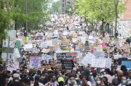 A large and diverse crowd of people fills a wide street lined with trees. Many hold protest signs with slogans such as “Know justice know peace,” “Black Lives Matter” and “Am I next?”