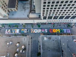An aerial photo of a downtown street between office buildings. The street has been painted with very large and colourful text reading “LA VIE DES NOIR.E.S COMPTE” with smaller white text reading “#BLACKLIVESMATTER.”