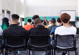 A group of young people in a classroom, seen from behind. They have a variety of skin colours, hair styles and clothes. 