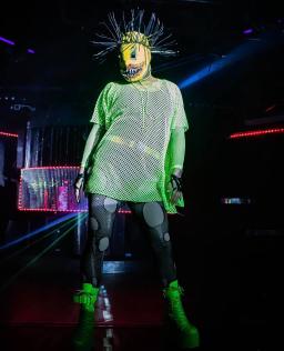 A drag queen wearing a yellow mask with sharp teeth, bright green mesh top and boots, and dark grey tights with large light grey circles.