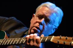 A close-up of a man with glasses and white hair playing a guitar.