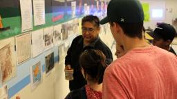 A man wearing glasses and holding a mug speaks to a small group of younger people. They are looking at a series of displays, including colourful images, maps, and text that cover one long wall of a large room.