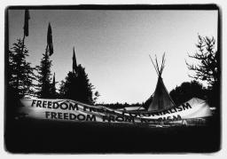 Black and white photo showing a large banner reading “Freedom from colonialism / Freedom from racism” set up in front of three flags and a teepee, in a forested landscape.