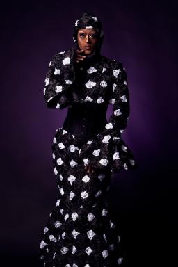A drag queen dressed from head to toe in a long, black mermaid style dress decorated with a white printed design.