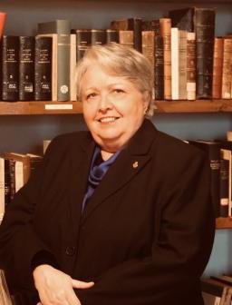 Randi Gage is an older woman with short, fair hair wearing a black blazer. She sits in front of a bookcase.