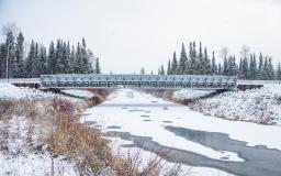 A metal bridge spans a river-like channel. The water is largely frozen, snow is on the ground and in the background are tall evergreen trees.