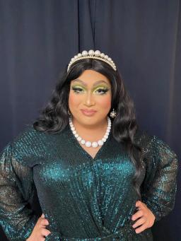 A drag queen wearing a shimmery, dark green dress, pearl tiara and white beaded necklace.