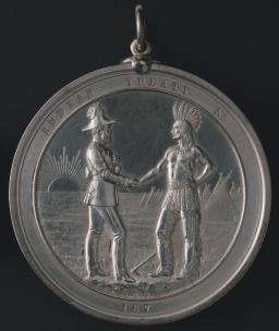 A round silver-coloured medal reading “INDIAN TREATY No.” along the top and “187” at the bottom. The large central section shows a distant sunrise and some teepees behind two figures shaking hands. On the left is a man dressed in formal colonial military clothing, and on the right a shirtless man wearing fringed pants and a feather headdress.