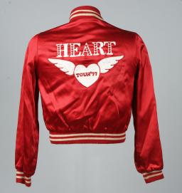 The back of a bright red satin jacket with the white text “HEART” in large capital letters above a white winged heart with “TOUR ‘77” written across it.