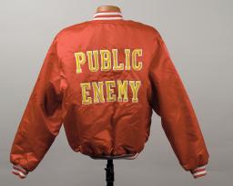 The back of a deep orangey-red bomber jacket decorated with “Public Enemy” in large yellow capital letters.