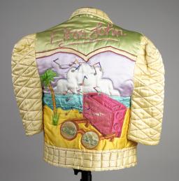 The back of a shiny yellow, quilted satin jacket decorated with the words “Elton John” above a beach scene with a purple sky, large white cloud, blue sea, dark yellow sand, a palm tree, a dark pink radio with music notes flying out of it, and a pair of round sunglasses in the foreground.