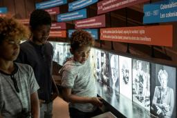 Three children look at a museum exhibit. The exhibit shows a series of black and white photos presented in a long row along a wall. Text panels of various sizes and colours are suspended above the photos.