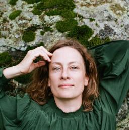 A woman with reddish hair and green eyes wearing an emerald green shirt, lies on a lichen-covered rock. One hand is behind her head; the other is touching her forehead.