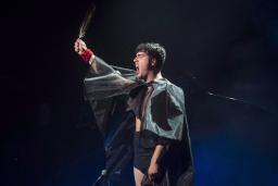 A person with short hair singing, while wearing a black cape and holding up a feather. 