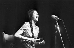 A woman with long braided hair sitting on a chair and singing into a microphone while playing a small hand rattle. 
