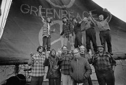 Eleven people posing in two rows in front of a boat’s mast and sail, gesturing with fists and peace signs. Five of them stand on the large spar holding the bottom of the sail, which is decorated with “GREENPEACE” in large letters above a peace sign.