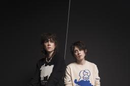 Two people with medium-long hair pose against a black backdrop. One wears a black sweater with a white sheep on the front, the other wears a white sweater with a blue mermaid.