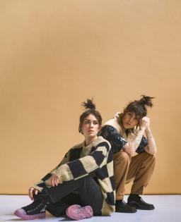 Two people with long hair in high ponytails pose against a beige backdrop and sit on a pale lavender coloured floor. 