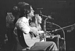Two people sit on chairs playing guitars and singing into microphones, with some other people mostly obscured behind them. 