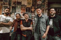 Group of five men in a band standing intentionally with a smile in front of a wall with band posters.