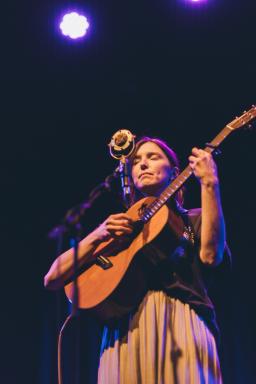 A woman playing guitar with emotion on a dark stage. She's standng behind a microphone with her eyes closed.