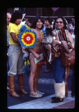 A photo showing three people in the foreground with a crowd in the background. On the right, a person with sunglasses and feathers in their hair wears a striped top, jeans and furry mukluks, holds an eagle feather fan and smiles at the camera. The person in the centre wears only moccasins, underwear and a few pieces of colourful, feathered fancy dance powwow regalia on his shoulders and back. Behind him, on the left, stands a tall person in a yellow t-shirt with green pants tucked into leather riding boots.