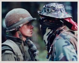 A uniformed soldier wearing a helmet stands in a tense face-to-face confrontation with someone wearing a camouflage jacket, hat and bandana covering their face. 