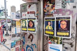 An exterior wall covered with graffiti and posters showing the faces of children and youth with text. A city landscape in what looks like a marketplace is in the background.