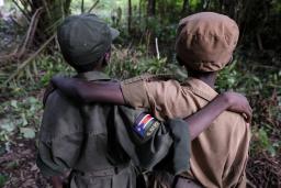 Two children seen from the back with their arms resting on each other’s shoulders. The one on the left is wearing an army green uniform with a South Sudan badge on the right arm and the other is wearing a tan uniform. Both are wearing hats that match their uniforms.