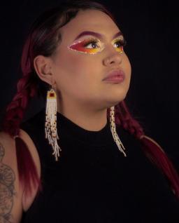 A person with long auburn hair, fiery eyeshadow and long white beaded earrings. They look proud and their eyeline is up.