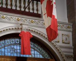 A bright red dress hangs in front of a rounded window in a large building that features ornate plasterwork accented with gold. A Canadian maple leaf flag hangs nearby.