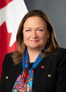 A headshot of Marie-Geneviève Mounier, a white woman with shoulder-length brown hair wearing a dark suit, a brightly coloured scarf of an Indigenous art print and a Canadian maple leaf pin.