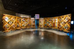 A large, curved freestanding wooden wall on which is mounted numerous small objects arranged in diamond and rectangular patterns. There is an open door in the centre of the wall.