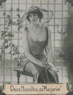 A black-and-white photo of a very convincing drag queen dressed in flapper-style clothes with a big summer hat, sitting on a wooden chair with a lattice behind her. There are vines growing up it in an elegant fashion. Under the photo, it says “Ross Hamilton as ‘Marjorie.’” 