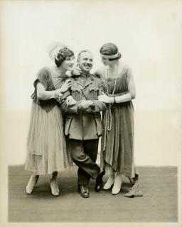 A black-and-white photo of two men dressed up as 1920s flapper girls. They are smiling adoringly and holding the arms of the uniformed officer who stands between them.