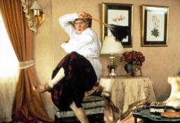 A man dressed in drag as a middle-aged woman, dancing with a feather duster and in a 1980s-style home. Her knee-high stockings are creeping down and she looks overwhelmed with the task of cleaning