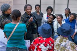 A group of women of all ages cluster around a woman at the centre who is making an emphatic point.