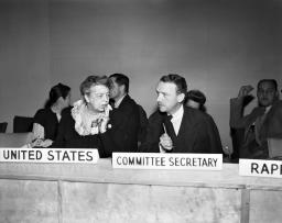 A woman, Eleanor Roosevelt, sits at a dais listening intently to the man on her left, John Peters Humphrey. In front of her is a plaque that reads “United States.” In front of him, a plaque reads “Committee Secretary.”