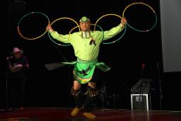 A hoop dancer dressed in a long-sleeve yellow top and green skirt is holding five hoops with arms outstretched like an eagle.