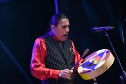 An Indigenous performer wearing a red, long-sleeve top and black, patterned vest is chanting and drumming.