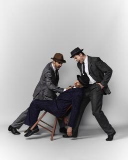 Two men dressed in dark suits and hats stand on either side of a similarly dressed man seated in a chair. They are pushing him and the chair backwards, grabbing him by his jacket and hollering at him in anger. The seated man's face expresses distress and fear.