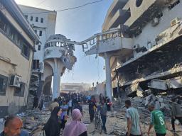 A group of men and women mull outside a thoroughfare between two buildings, surveying the damage following damage sustained in military conflict.