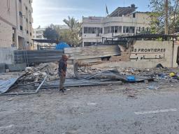 A man wearing a grey sweatshirt and grey pants standing outside of the damaged wall of a Doctors Without Borders/Médecins Sans Frontières building amidst debris and rubble.